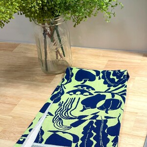 May include: A folded kitchen towel with a vibrant pattern of navy blue shapes on a light green background. The towel has a white and gray striped border. A glass jar with green plants is in the background.