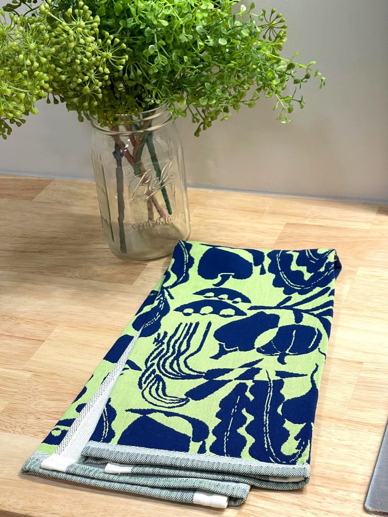 May include: A folded kitchen towel with a vibrant pattern of navy blue shapes on a light green background. The towel has a white and gray striped border. A glass jar with green plants is in the background.