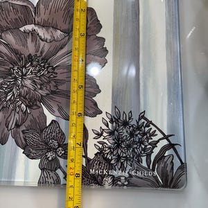 Puede incluir: Una bandeja rectangular de vidrio con un dise&ntilde;o floral en tonos grises y blancos. La bandeja mide aproximadamente 23 cm de largo. La marca "MACKENZIE-CHILDS" est&aacute; impresa en la bandeja.