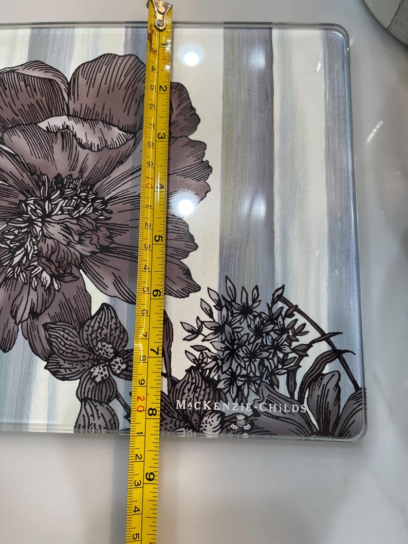 Puede incluir: Una bandeja rectangular de vidrio con un dise&ntilde;o floral en tonos grises y blancos. La bandeja mide aproximadamente 23 cm de largo. La marca "MACKENZIE-CHILDS" est&aacute; impresa en la bandeja.