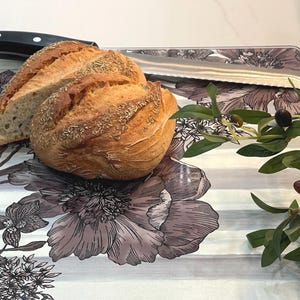 Puede incluir: Una hogaza de pan, parcialmente rebanada, descansa sobre una tabla de cortar con estampado floral con un cuchillo de acero inoxidable. Una rama de olivo con aceitunas est&aacute; a la derecha. La tabla tiene un fondo a rayas grises y blancas.
