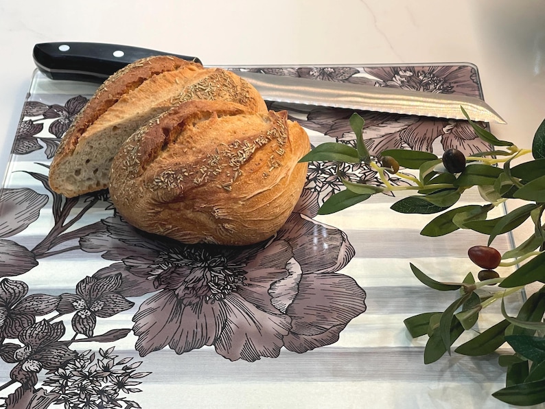 Puede incluir: Una hogaza de pan, parcialmente rebanada, descansa sobre una tabla de cortar con estampado floral con un cuchillo de acero inoxidable. Una rama de olivo con aceitunas est&aacute; a la derecha. La tabla tiene un fondo a rayas grises y blancas.