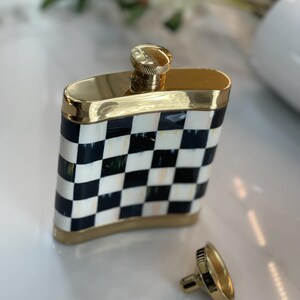 May include: A gold-trimmed flask with a black and white checkered pattern. The flask has a gold cap and a separate, matching cap. The flask is rectangular and has a shiny finish.