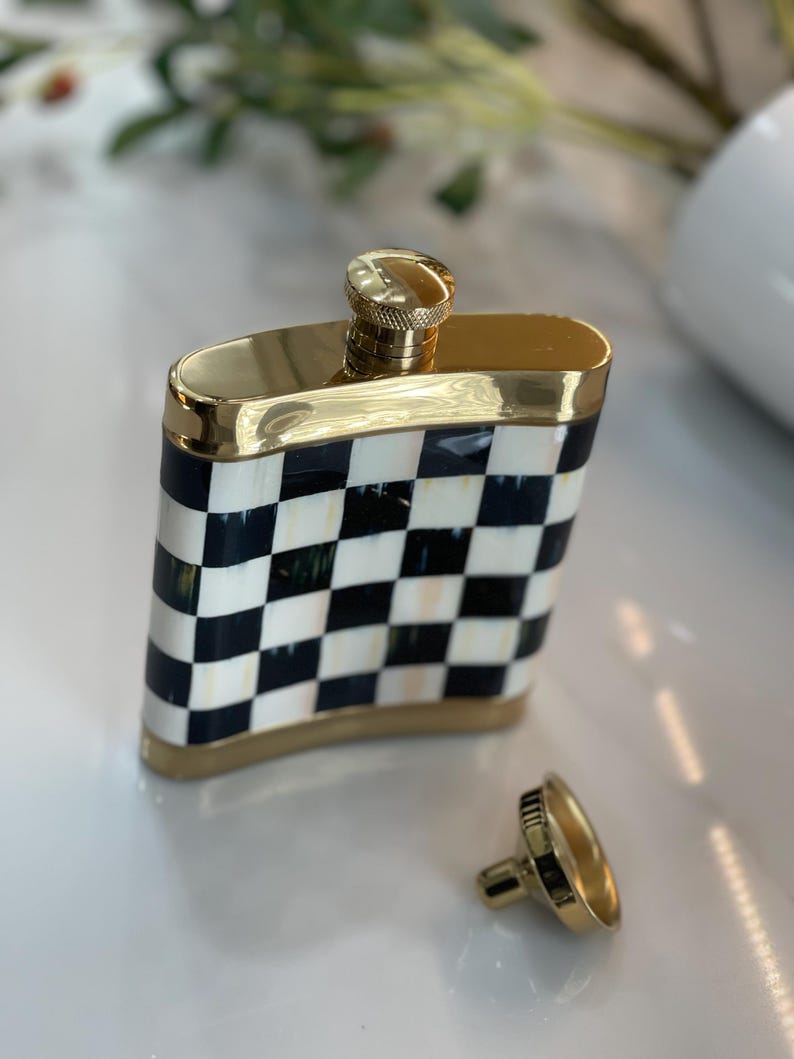 May include: A gold-trimmed flask with a black and white checkered pattern. The flask has a gold cap and a separate, matching cap. The flask is rectangular and has a shiny finish.
