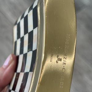 May include: A decorative flask with a gold-colored metal body and a black and white checkered pattern on the front. The flask has rounded edges and the text "MacKenzie-Childs" is visible on the side. The flask is made in India.