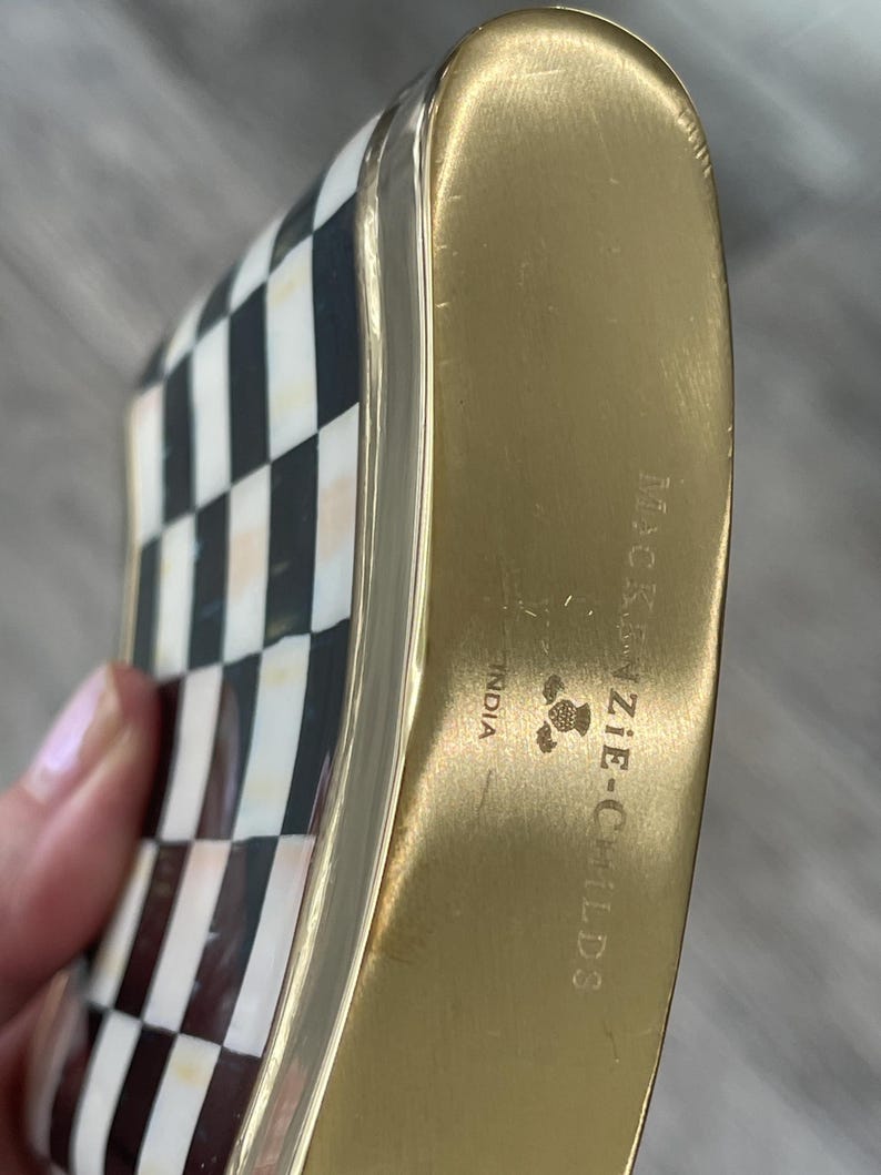 May include: A decorative flask with a gold-colored metal body and a black and white checkered pattern on the front. The flask has rounded edges and the text "MacKenzie-Childs" is visible on the side. The flask is made in India.