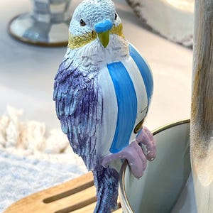 以下が含まれることがあります： 青、白、紫の配色が施された、装飾用のセラミック製のセキセイインコの置物。鳥は金属製の容器の端に止まっています。この置物は、羽の模様が細かく、黄色い嘴が特徴です。