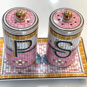 Puede incluir: Un juego decorativo de salero y pimentero con un dise&ntilde;o de mosaico rosa, dorado y blanco. El juego incluye dos saleros cil&iacute;ndricos con tapas doradas y una bandeja rectangular. Las letras "P" y "S" se muestran.