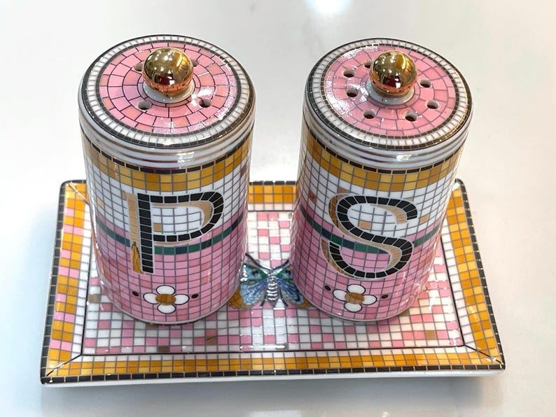Puede incluir: Un juego decorativo de salero y pimentero con un dise&ntilde;o de mosaico rosa, dorado y blanco. El juego incluye dos saleros cil&iacute;ndricos con tapas doradas y una bandeja rectangular. Las letras "P" y "S" se muestran.