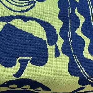 May include: Close-up of a folded textile with a repeating pattern. The design features large, abstract shapes in navy blue against a light green background. The fabric appears to be woven.
