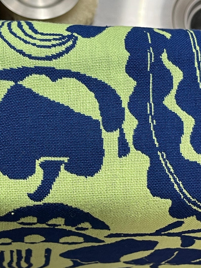 May include: Close-up of a folded textile with a repeating pattern. The design features large, abstract shapes in navy blue against a light green background. The fabric appears to be woven.
