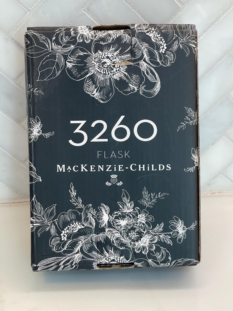 May include: A dark gray flask box with white floral illustrations. The number "3260" is centered in white, with "FLASK" and "MACKENZIE-CHILDS" below. The box has a decorative floral design.