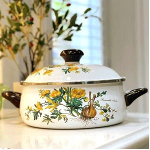 May include: White enamel cooking pot with a lid, featuring a floral and botanical design in yellow and green. The pot has brown handles and a black knob on the lid. The design includes a botanical illustration of a bulb.