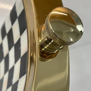 May include: A gold-colored flask with a checkered black and white pattern on one side. The flask has a shiny, smooth finish and a gold-colored screw-on cap. The flask is designed to hold liquids.