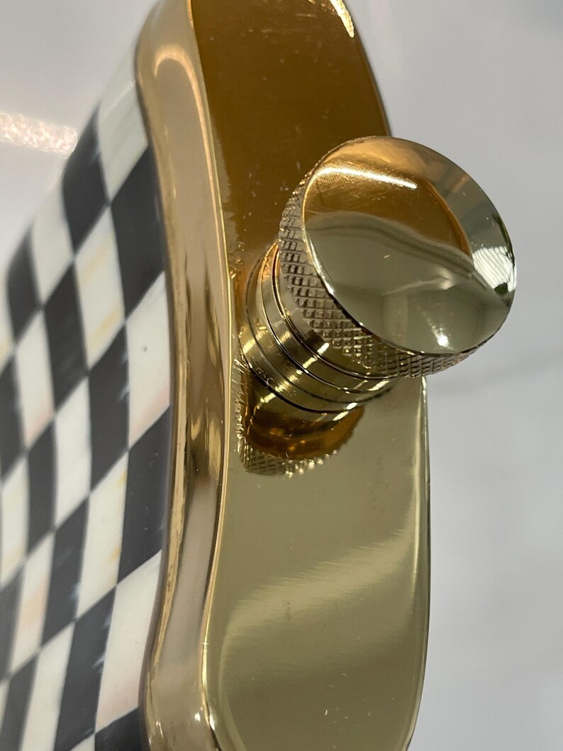 May include: A gold-colored flask with a checkered black and white pattern on one side. The flask has a shiny, smooth finish and a gold-colored screw-on cap. The flask is designed to hold liquids.