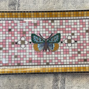 Puede incluir: Bandeja decorativa rectangular con dise&ntilde;o de mosaico. La bandeja presenta un patr&oacute;n de cuadros rosas y blancos con una mariposa en el centro. La mariposa tiene alas azules, verdes y amarillas. El borde es dorado y negro.