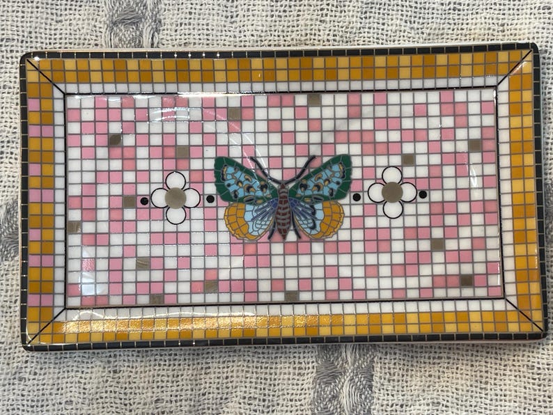 Puede incluir: Bandeja decorativa rectangular con dise&ntilde;o de mosaico. La bandeja presenta un patr&oacute;n de cuadros rosas y blancos con una mariposa en el centro. La mariposa tiene alas azules, verdes y amarillas. El borde es dorado y negro.
