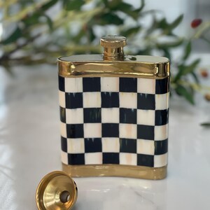 May include: A decorative flask with a gold-colored top and trim. The flask features a black and white checkered pattern. A gold-colored funnel sits next to the flask.