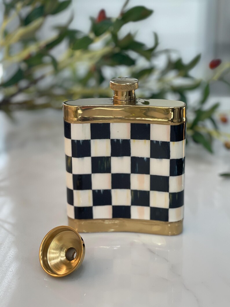 May include: A decorative flask with a gold-colored top and trim. The flask features a black and white checkered pattern. A gold-colored funnel sits next to the flask.