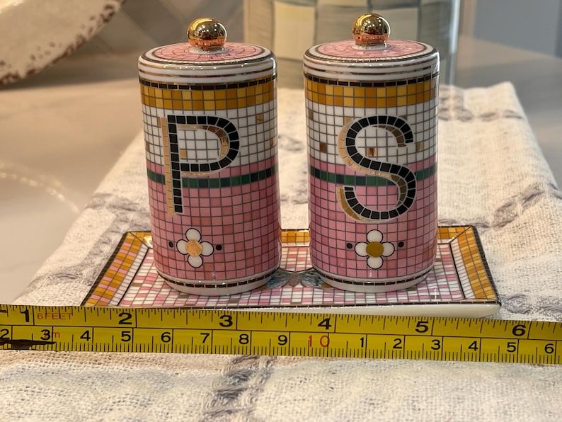 Puede incluir: Un juego de salero y pimentero de cer&aacute;mica con dise&ntilde;o de mosaico rosa, dorado y blanco. Los saleros y pimenteros tienen tapas doradas y est&aacute;n etiquetados con las letras "P" y "S". El juego se encuentra sobre una bandeja a juego.