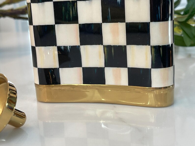May include: A decorative ceramic container with a black and white checkered pattern. The base of the container is a gold color. The container is rectangular and has a smooth, glossy finish.
