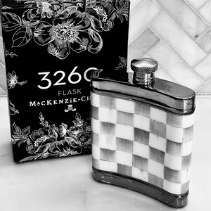 May include: A black and white flask with a checkered pattern and a silver cap. The flask is next to a black box with floral designs and the text "3260 FLASK MACKENZIE-CH". The flask is a stylish accessory.