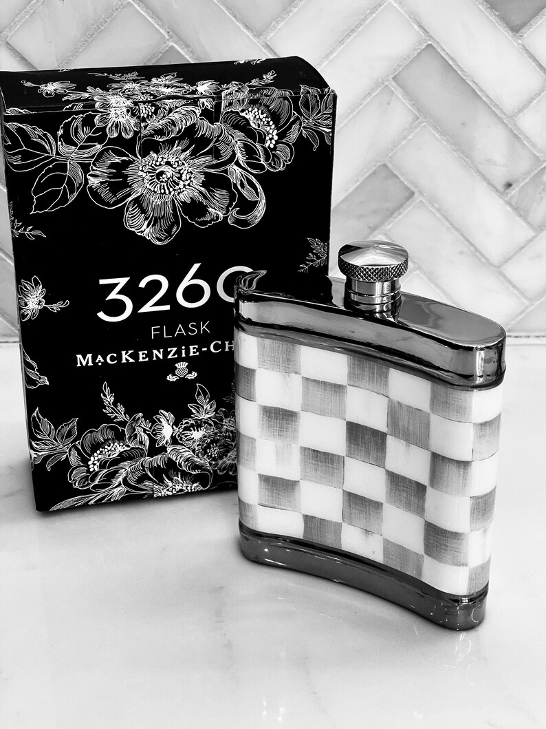 May include: A black and white flask with a checkered pattern and a silver cap. The flask is next to a black box with floral designs and the text "3260 FLASK MACKENZIE-CH". The flask is a stylish accessory.