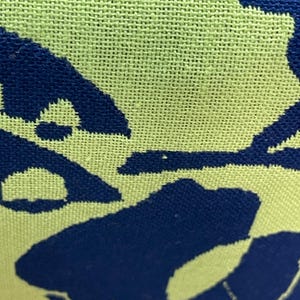 May include: Close-up of a woven fabric with a light green background and a dark blue pattern. The pattern appears to be a stylized floral or abstract design, with the blue shapes contrasting against the green.