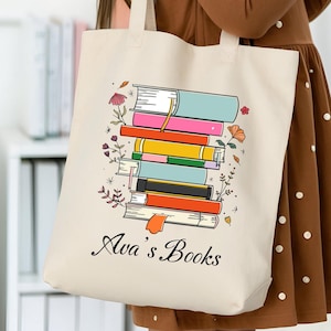 May include: A beige tote bag featuring a stack of colorful books with floral accents. The text "Ava's Books" is printed below the books. The bag is held by a young girl wearing a brown dress.