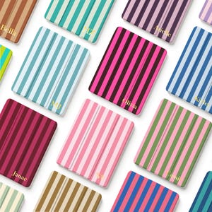 Könnte beinhalten: Eine Sammlung rechteckiger Tablet-Hüllen mit vertikalen Streifen in verschiedenen Farbkombinationen wie Pink, Blau, Grün und Braun. Jede Hülle ist mit einem goldfarbenen Namen oder Initialen personalisiert, die in der Ecke aufgedruckt sind.
