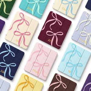 May include: A collection of rectangular tablet cases featuring a textured fabric finish and a printed bow pattern. Each case displays a unique solid color, including burgundy, navy, pink, and yellow, with personalized names or initials printed in gold.