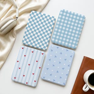 May include: Four rectangular e-reader cases featuring distinct patterns: one with a blue and white checkerboard, one with a blue and white gingham plaid, one with light blue stripes and small red hearts, and one with blue bows and white dots.