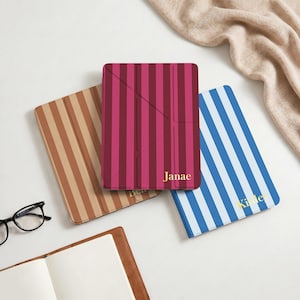 Könnte beinhalten: Drei rechteckige Tablet-Hüllen mit vertikalen Streifen in Hellbraun, Dunkelmagenta und Blau. Die magentafarbene Hülle ist mit dem Namen Janae in Gold personalisiert, während die blaue Hülle den Namen Kinley in Gold trägt. Eine schwarze Brille liegt daneben.