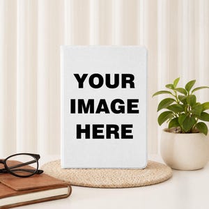 May include: A white rectangular tablet case or notebook cover featuring the black text YOUR IMAGE HERE centred on the front surface. The object stands upright on a round woven mat, positioned next to a brown leather-bound journal and glasses.