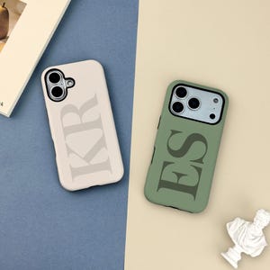 May include: Two phone cases are shown, one cream and one olive green. The cream case features the letters "KR" in a darker tone, while the green case displays "ES". Both cases have a camera cutout and are on a blue and beige surface.