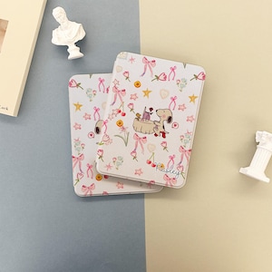 Puede incluir: Dos estuches decorativos blancos con un patrón repetido de lazos rosas, flores, estrellas y un perro de dibujos animados. La palabra "Paisley" está escrita en cursiva en la esquina inferior derecha del estuche.