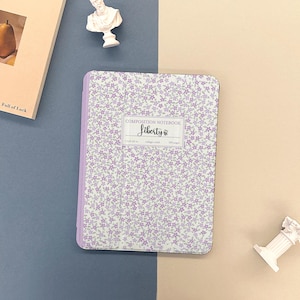 Puede incluir: Un cuaderno de composición con un estampado floral en tonos morados y blancos. El cuaderno tiene un lomo morado claro y el texto "Liberty" en la portada. El cuaderno mide 18,4 x 13,3 cm y contiene 100 páginas.