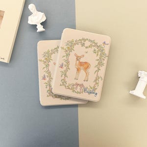 May include: Two rectangular, off-white boxes with a floral and deer design. The boxes feature a light brown deer in the center, surrounded by a floral wreath. The word "Paisley" is written in cursive at the bottom.