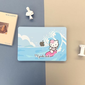 May include: A light blue laptop skin featuring a cartoon character surfing on a pink surfboard in front of a wave. The design includes an Apple logo and the name "Gabriel".