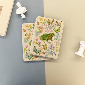 May include: Two rectangular cream-colored cases with a floral and butterfly design, featuring a green frog. The cases have a watercolor style with the word "Paisley" in script. The background is a mix of blue and beige.