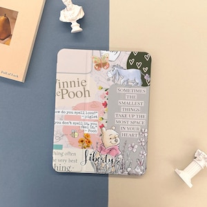 Può includere: Una custodia decorativa per tablet con personaggi e citazioni di Winnie the Pooh. La custodia ha uno schema di colori floreali e pastello con illustrazioni di Pooh, Eeyore, una farfalla e un'ape. Il testo include "Liberty" e citazioni sull'amore.
