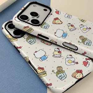 May include: Two white phone cases with a repeating pattern of Hello Kitty characters, food items, and small red hearts. The cases have a glossy finish and black edges. The phone's camera lenses are visible.