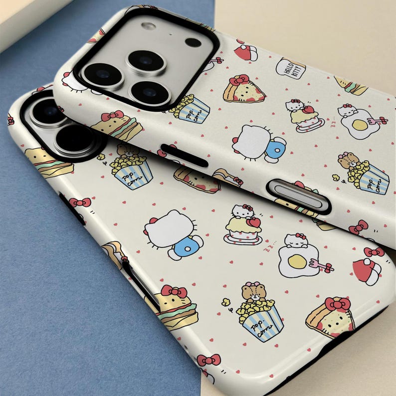 May include: Two white phone cases with a repeating pattern of Hello Kitty characters, food items, and small red hearts. The cases have a glossy finish and black edges. The phone's camera lenses are visible.