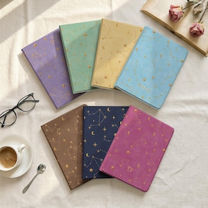 May include: A collection of seven rectangular notebooks in purple, light green, yellow, light blue, brown, dark blue, and magenta, each featuring a gold foil celestial pattern of stars, moons, and constellations printed on the fabric-textured covers.