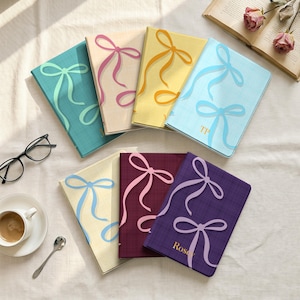 May include: A collection of seven rectangular notebooks with textured covers, each featuring a large printed bow graphic. The notebooks come in teal, beige, yellow, light blue, cream, maroon, and purple, with some displaying initials or names.