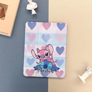 May include: Tablet case featuring a pink and blue cartoon character design. The case has a heart pattern in pink and blue. The characters are on a light blue and white striped background. The word "Liberty" is written on the case.