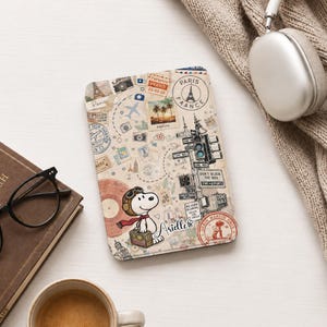 Può includere: Una custodia rettangolare per e-reader con un collage a tema viaggio che raffigura Snoopy con occhiali da aviatore e una sciarpa rossa, mentre tiene una valigia. Il design include francobolli vintage, un semaforo e il nome Arielle.