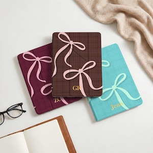 May include: Three rectangular tablet cases in burgundy, dark brown, and turquoise, each featuring a textured fabric-like finish and a light pink ribbon graphic. The brown case displays the gold text GR, and the turquoise case displays the gold text Jenn.