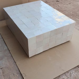 Op de afbeelding: Een rechthoekige salontafel, opgebouwd uit talrijke vierkante, gebroken witte tegels. De tafel staat op een stuk karton en toont een modern, geometrisch ontwerp. De tegels hebben een glanzende afwerking.