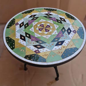 Op de afbeelding: Ronde mozaïektegel tafel met een geometrisch patroon in groen, geel, paars, zwart en wit. De tafel heeft een zwart metalen frame en poten. De tafel heeft een diameter van ongeveer 76 cm.